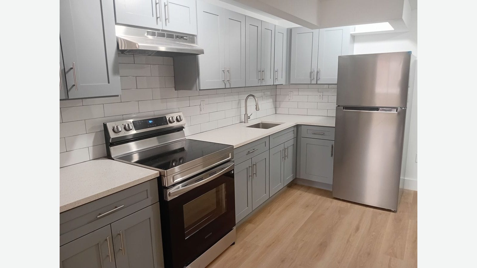 Photograph of a kitchen with gray shaker cabinets above and below, white subway tile, a white speckled counter with an undermount sink and arched faucet, a stainless electric range with a smooth glass top and a range hood, a stainless refrigerator at right and light wood-look flooring