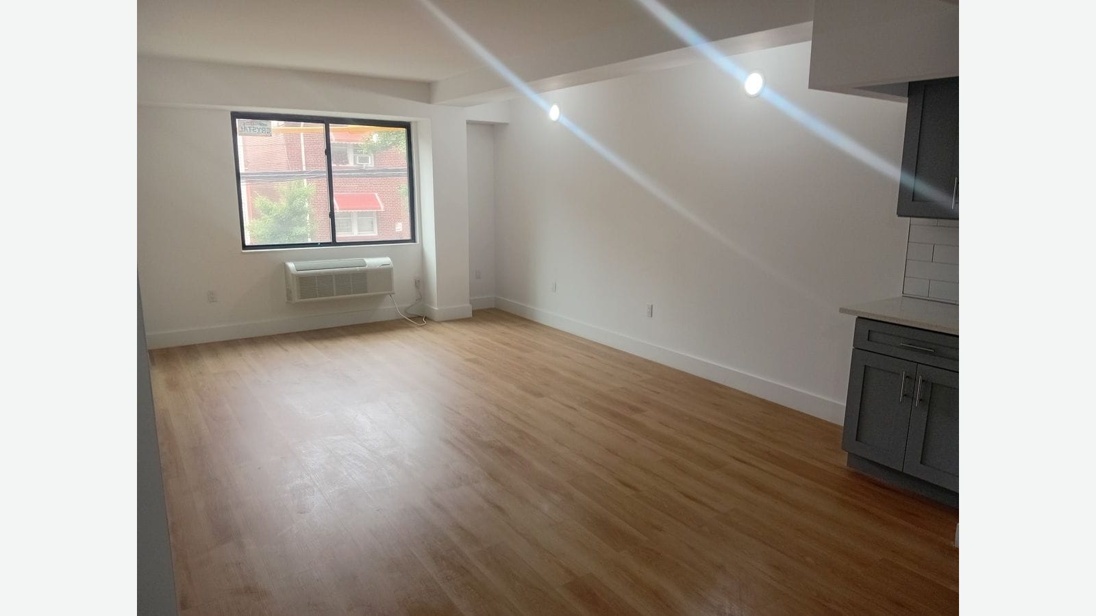 Photograph of an empty main room with light wood-look flooring, white walls, recessed ceiling lights, a heating and cooling unit under a dark-framed window looking onto a brick building, and gray kitchen cabinets and a counter at the right edge