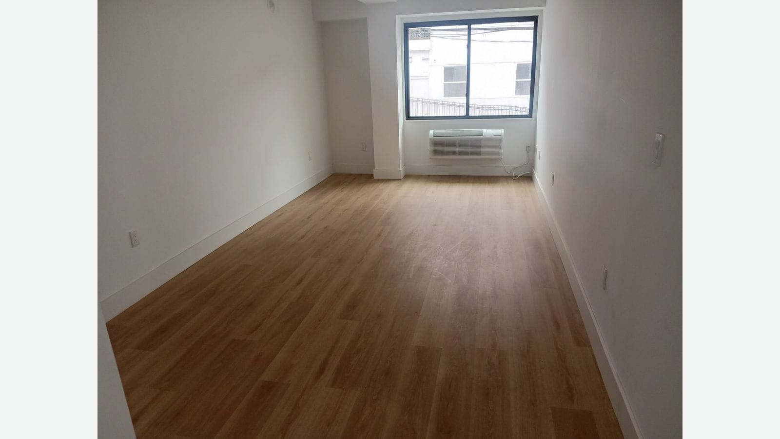 Photograph of a long, empty room with light wood-look flooring and white walls, ending at a dark-framed window with a heating and cooling unit below it