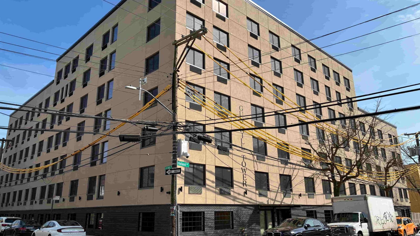 Photograph of a seven-story building with tan stucco walls above a dark gray brick base, seen from a street corner under a blue sky, with the words Gun Hill Tower in raised letters running down one wall, a utility pole with many black and yellow-sheathed wires in front, green street signs reading Holland Av and E 213 St, and parked cars and a white box truck along the curb
