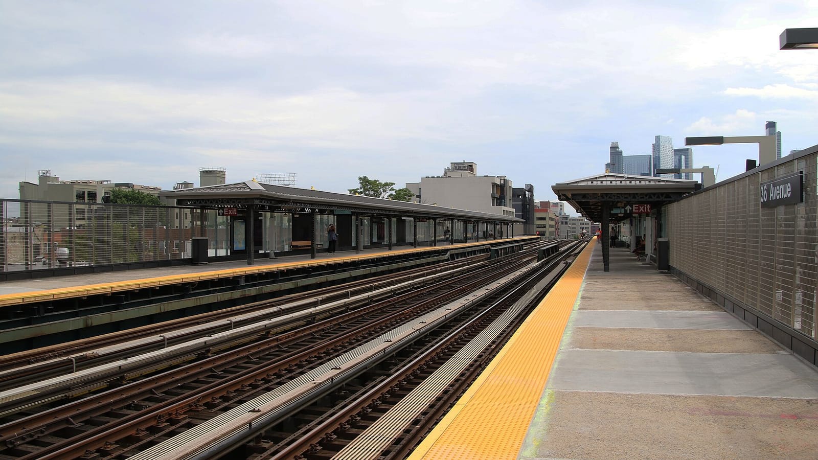 Photograph of an elevated subway station with two side platforms, a yellow platform edge, canopies marked Exit and a sign reading 36 Avenue, with low buildings and Long Island City towers behind under a cloudy sky