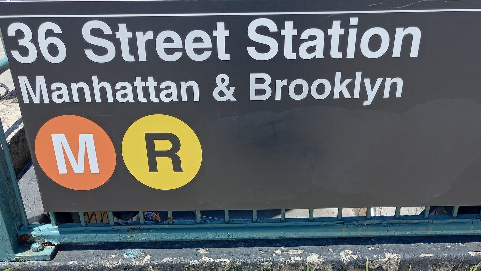 Photograph of a dark station sign reading 36 Street Station, Manhattan and Brooklyn, with orange M and yellow R route circles, on a green railing