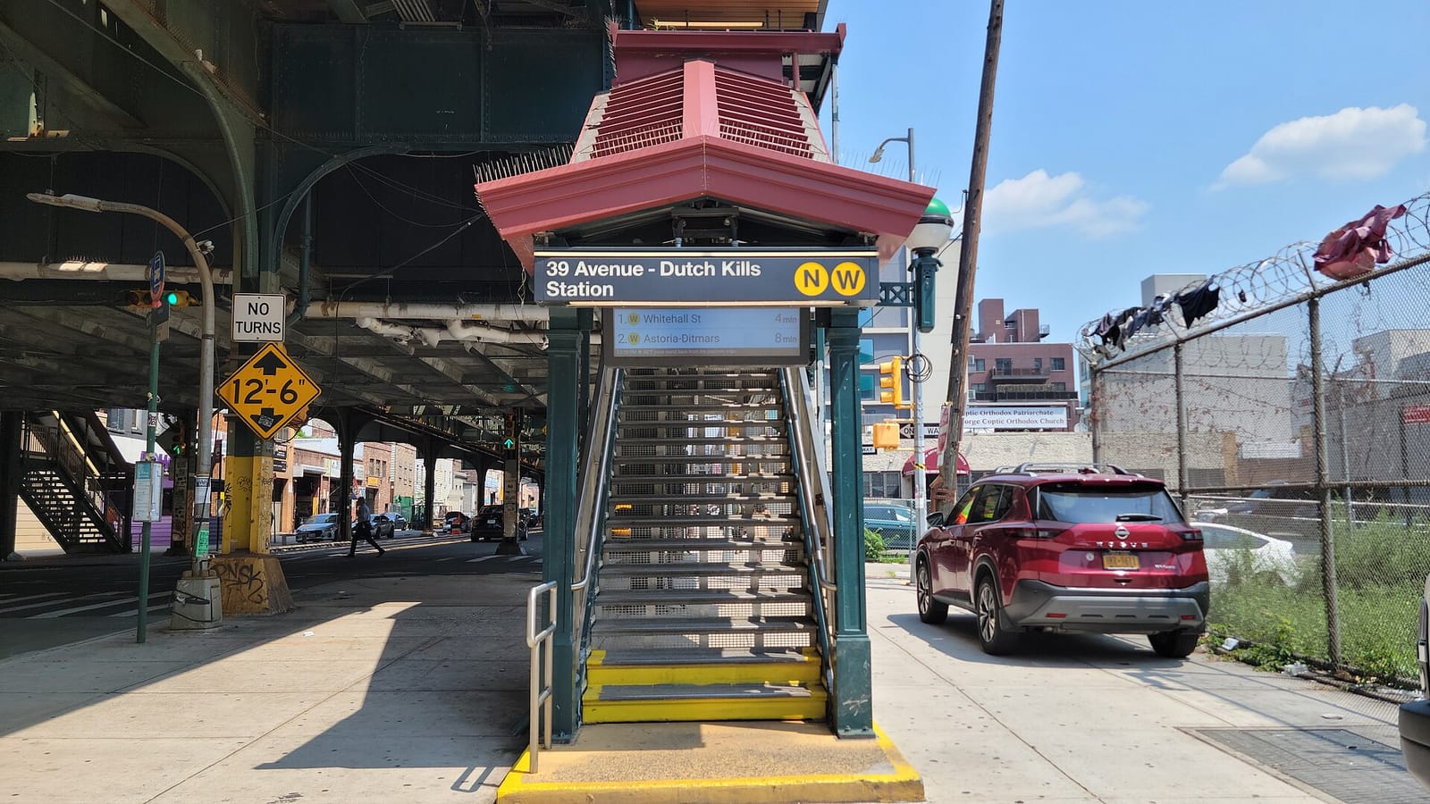 Photograph of a subway stair with a red pagoda-style canopy and a sign reading 39 Avenue - Dutch Kills Station with N and W symbols, under an elevated steel track, with a red SUV parked beside it and a chain-link fence topped with razor wire