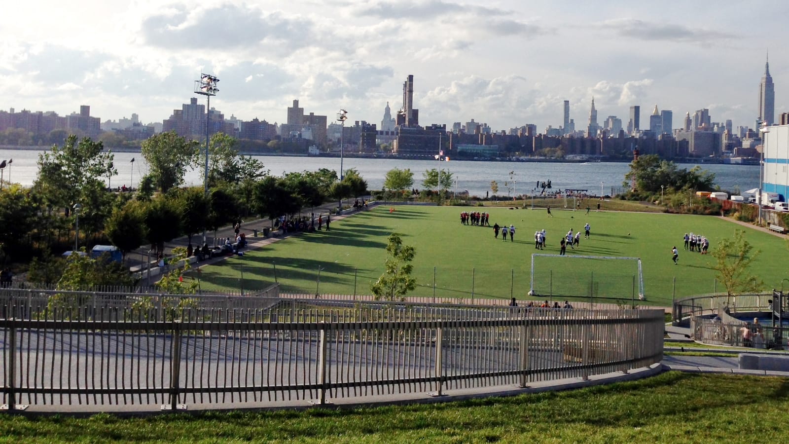 A large artificial-turf field with soccer goals and small groups of distant players, a curved metal fence in the foreground, trees, the East River and the Midtown Manhattan skyline beyond under scattered clouds