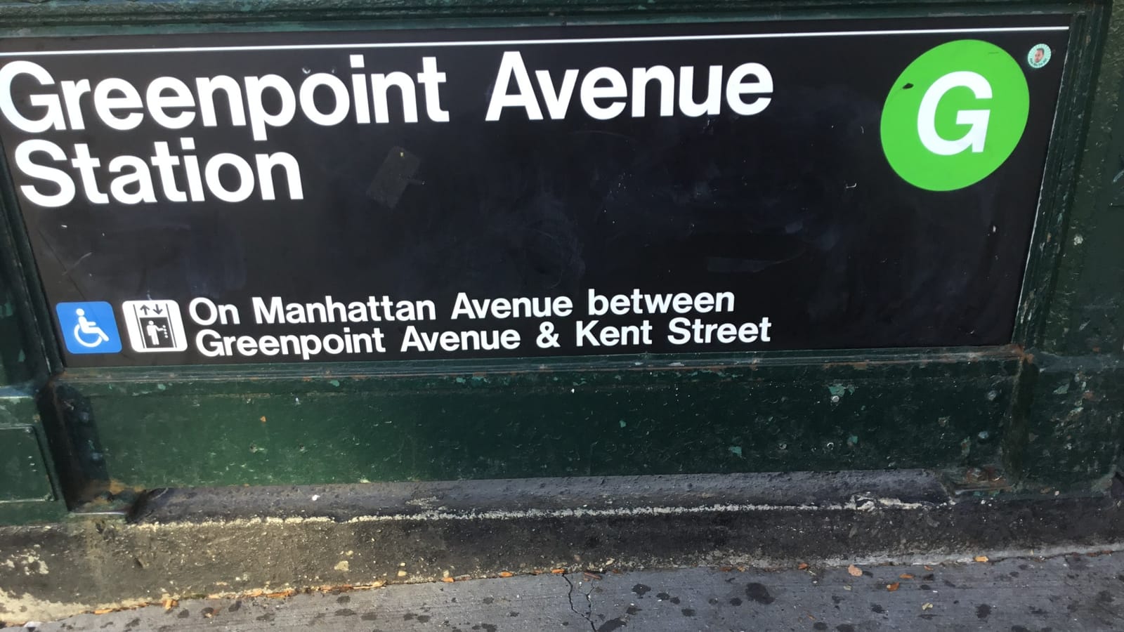 Close-up of a black subway entrance sign reading Greenpoint Avenue Station with a green G circle, and wheelchair and elevator symbols beside the words On Manhattan Avenue between Greenpoint Avenue and Kent Street, above a worn green railing base and sidewalk
