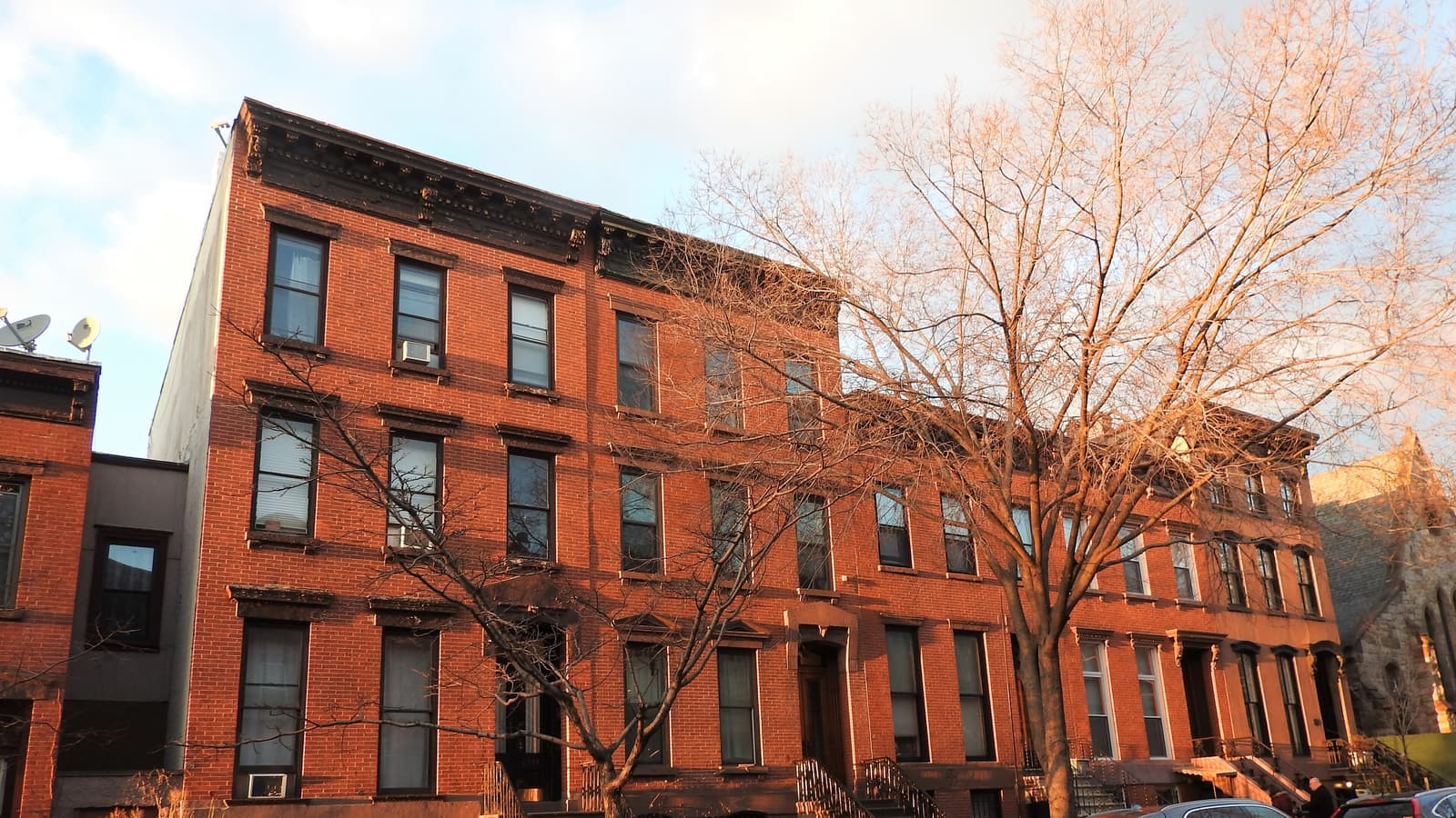 A row of four-story red brick row houses with stoops and dark cornices on Kent Street, lit orange by low sun, with bare trees in front and the tops of parked cars at the bottom edge