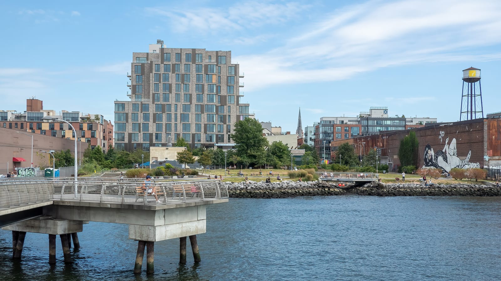 A concrete pier with metal railings over blue water in the foreground, a grassy waterfront park with trees and a rocky edge behind it, a 12-story apartment building, a water tower, a church spire and a large mural on a brick wall under a blue sky