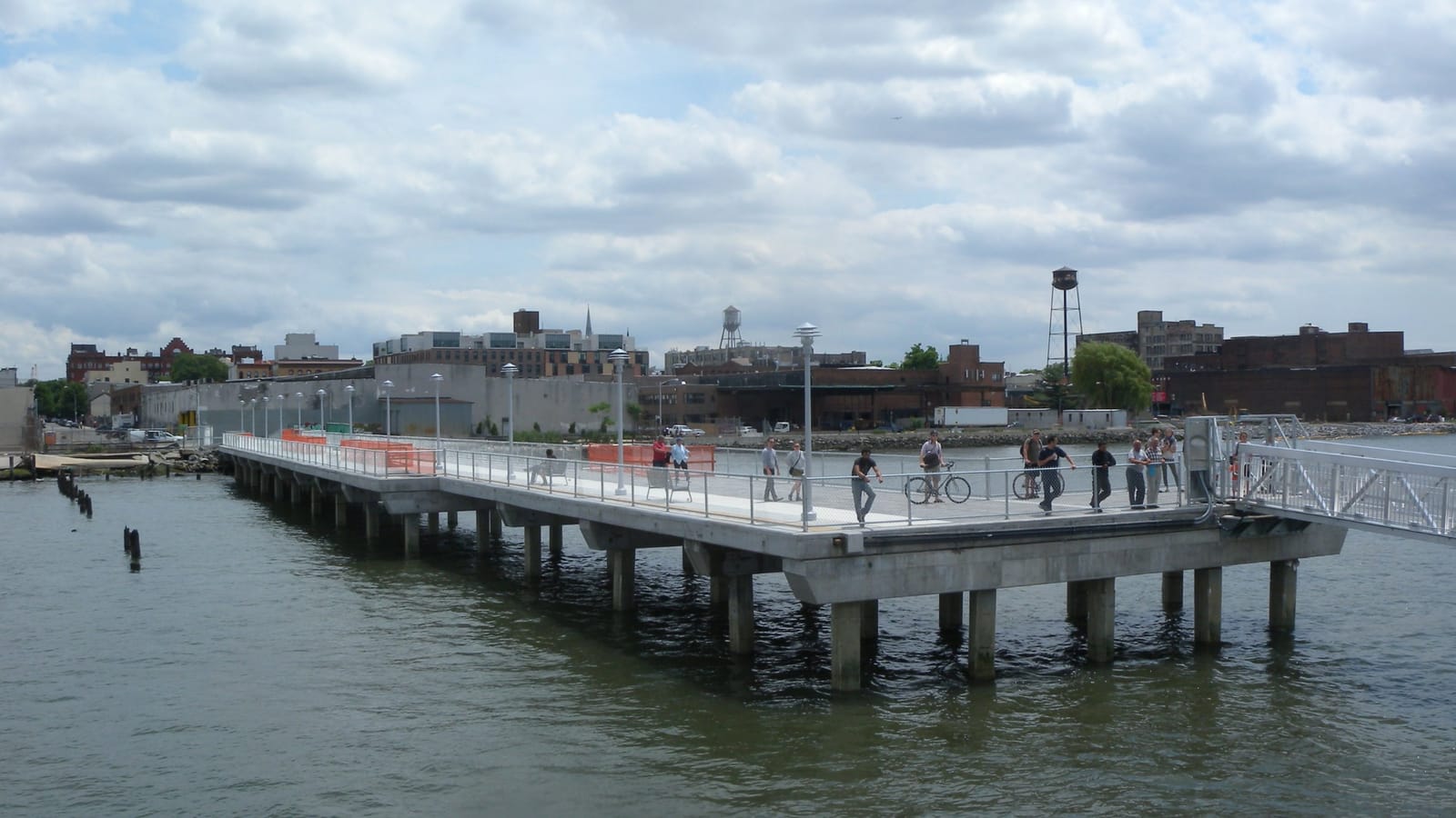 A long concrete pier on pilings over gray-green river water, with a few people walking and cycling on it far from the camera, and low brick warehouses, a water tower and a few taller buildings on the shore under a cloudy sky