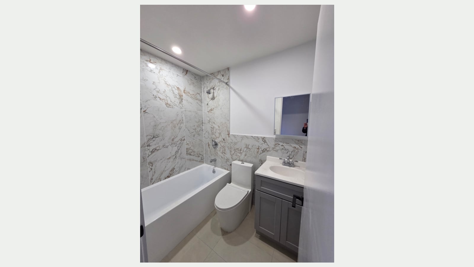 Photograph of a bathroom with marble-look wall tile around a white tub with a shower head and curtain rod, a white toilet, a gray vanity with a white sink, a small mirror cabinet and beige floor tile