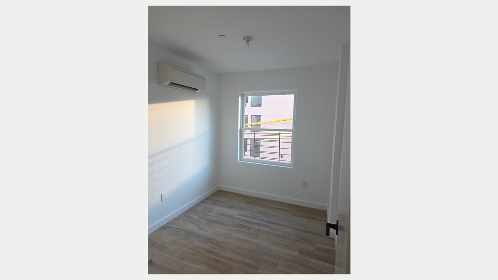 Photograph of an empty small room with wood-look plank floors, white walls, a wall-mounted split unit near the ceiling, sunlight on the left wall and a window looking out on a white building with railings and a yellow beam