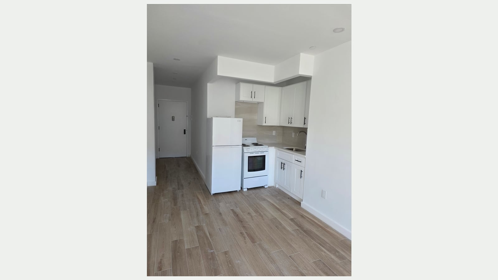 Photograph of an empty studio with wood-look plank floors, a white refrigerator, a white electric range with coil burners, white kitchen cabinets with black handles, a beige tile backsplash and a sink, with a hallway leading to the apartment door