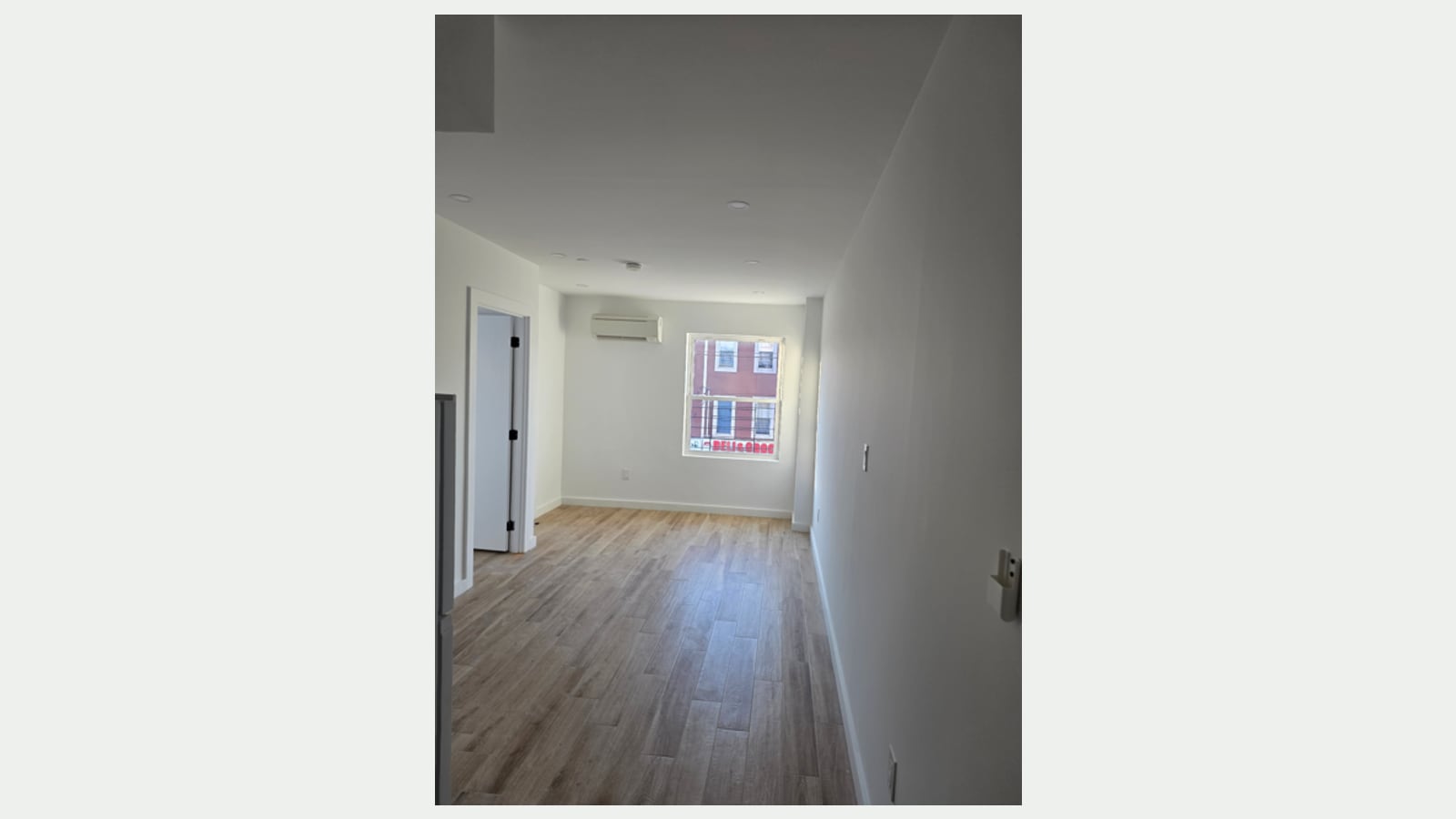 Photograph of an empty, narrow living room with wood-look plank floors, white walls, recessed ceiling lights, a wall-mounted split heating and cooling unit above a window, an open door to another room at left, and brick buildings and a red store sign seen through the window