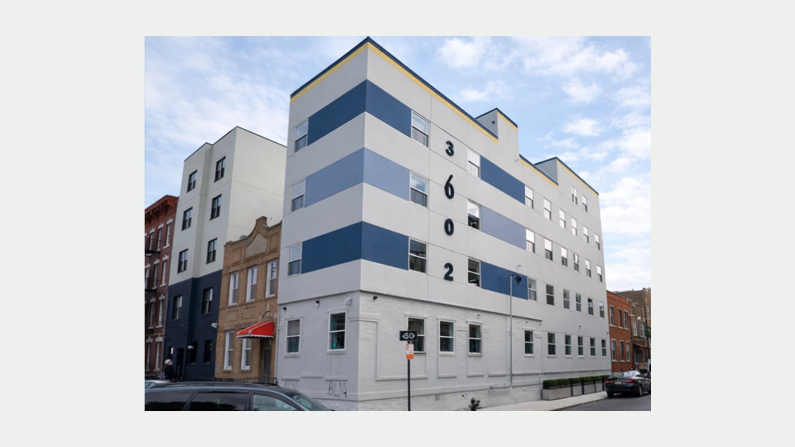 Photograph of a five-story corner apartment building painted light gray with wide dark and light blue bands across the upper floors, the large black numbers 3602 down its side, a painted brick ground floor with white-framed windows, a street sign on a pole, parked cars, and a white five-story building and a tan brick house next door under a partly cloudy sky