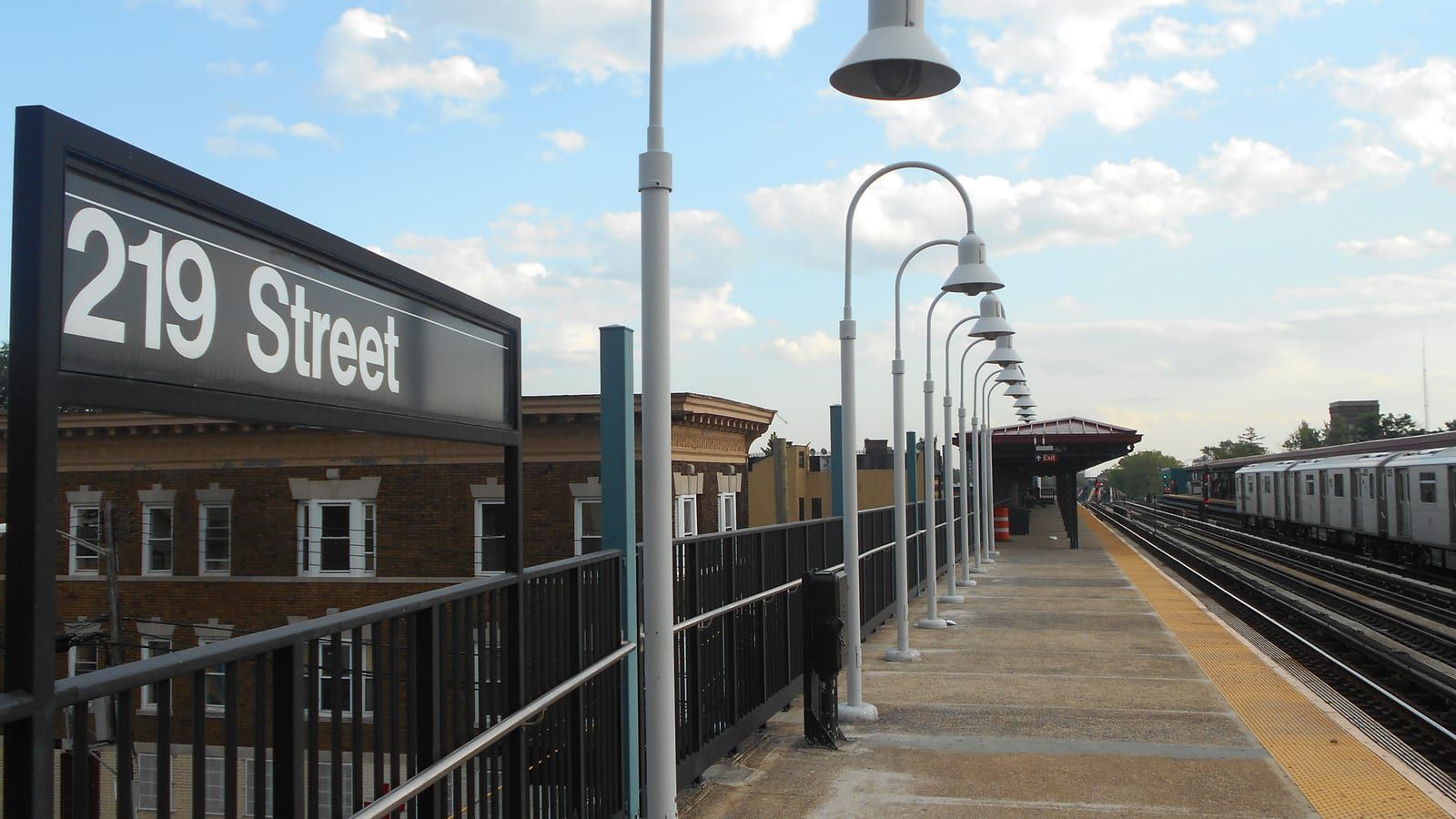 An open-air train platform with a large sign reading 219 Street at left, a black railing, a row of white curved lamp posts, a red-roofed shelter in the distance, a silver subway train on the far track at right and a brown brick apartment building below the platform
