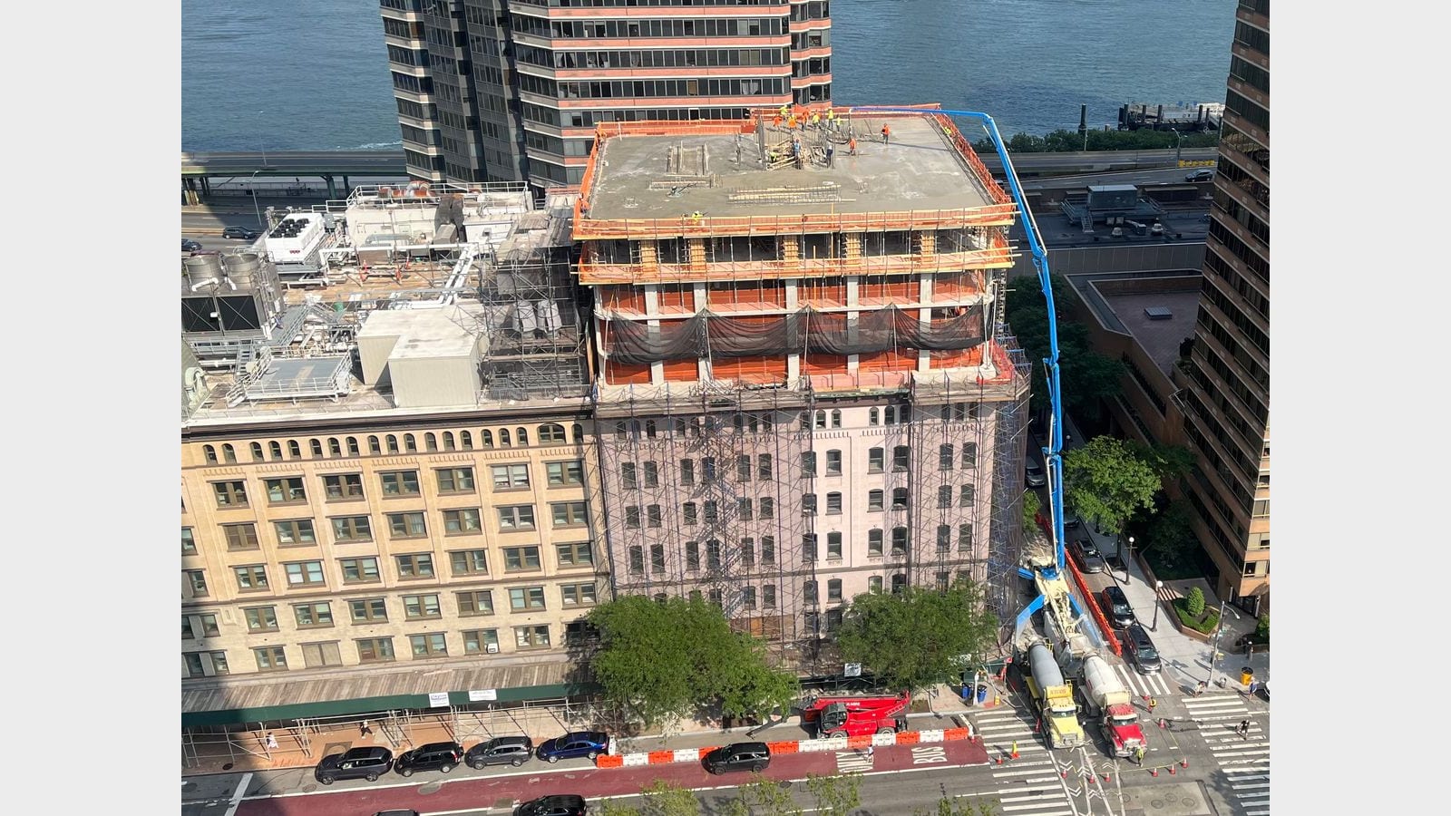 Photograph from a high window of 650 First Avenue during construction: new concrete floors with orange safety netting and workers on the roof slab rise above an older pink brick building wrapped in scaffolding, a blue concrete pump boom reaches up from trucks on First Avenue, an older tan building stands at left, and the East River and FDR Drive are behind
