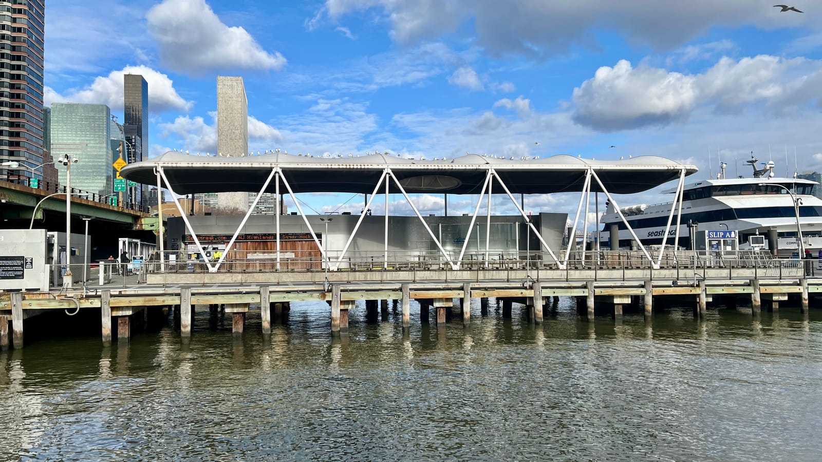 A ferry landing on wooden piles over the East River with a long white wavy canopy on angled white supports, a ferry docked at right beside a sign reading Slip A, a highway viaduct at left and office towers behind
