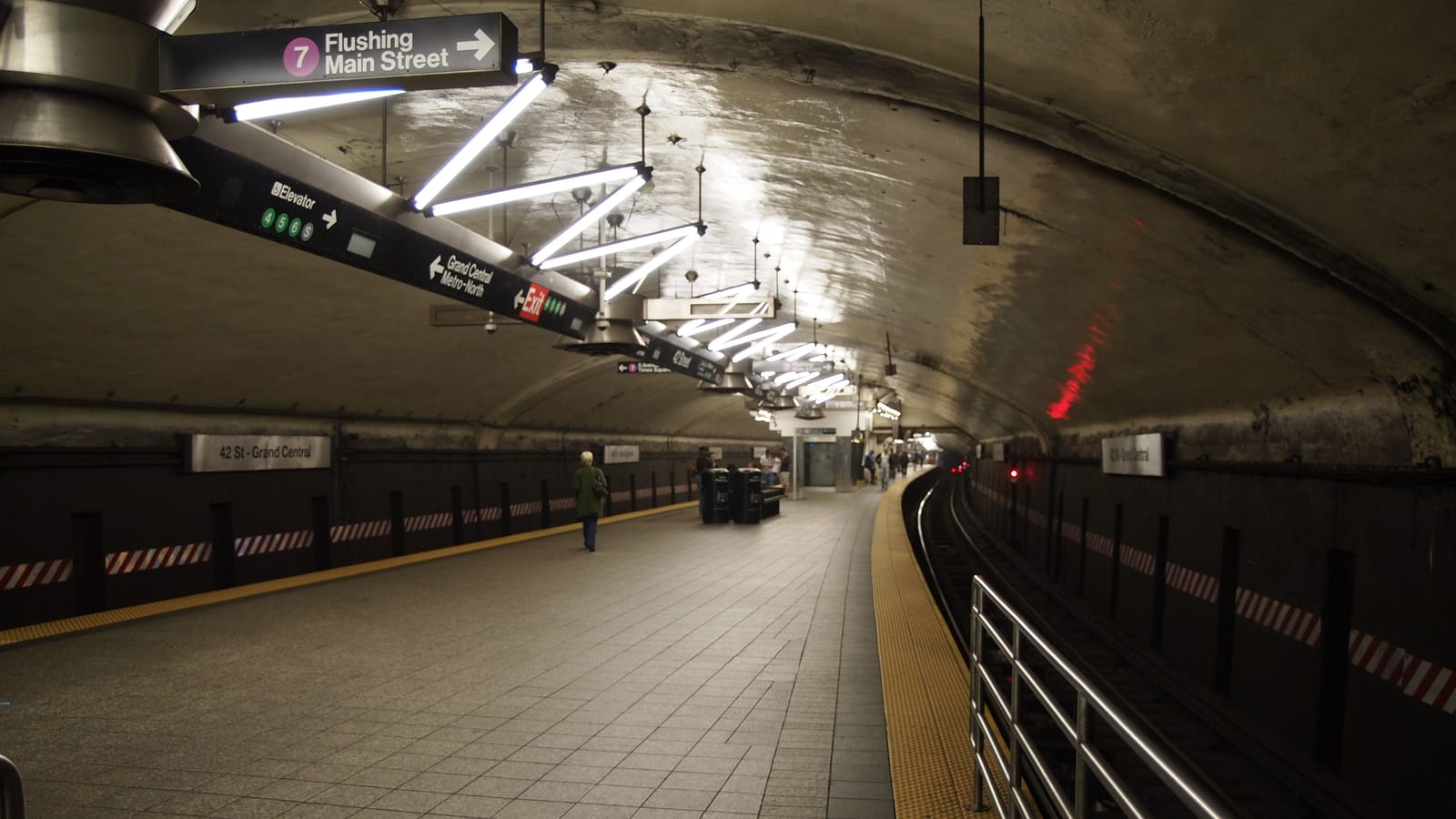 Underground subway platform under a curved tunnel ceiling, with a sign reading 7 Flushing Main Street, angled fluorescent lights, a 42 St - Grand Central name sign on the wall, a curving track at right behind a railing and a few people far down the platform