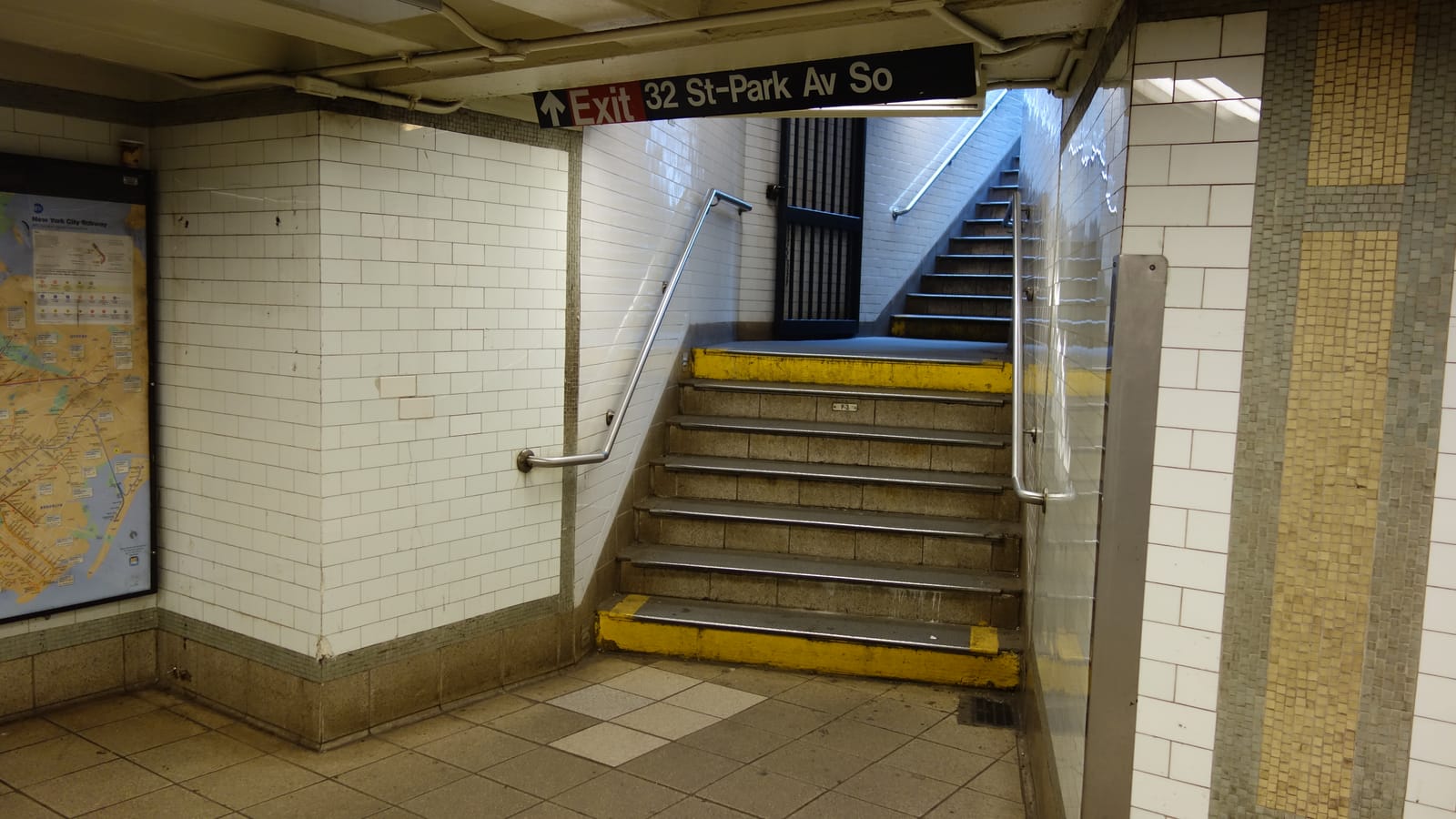 White-tiled subway passage with a stairway rising to street level under a sign reading Exit 32 St-Park Av So, yellow-edged steps, metal handrails and a subway map on the wall at left