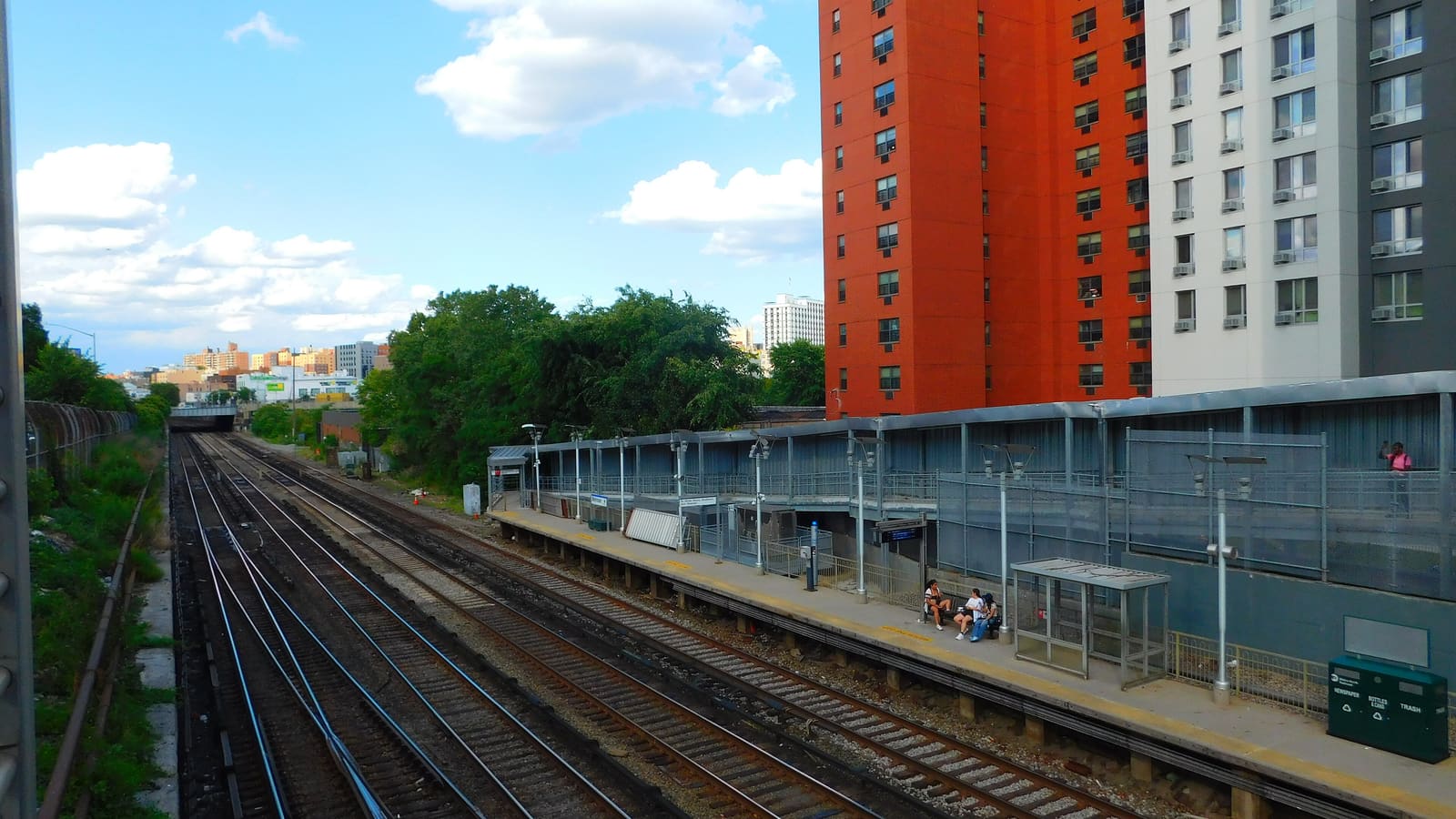 Photograph from above of a commuter rail station in a cut below street level, with four tracks at left, a long platform with a shelter, lamp posts and a few people sitting on a bench, a metal-clad station wall, and tall orange and white apartment towers on the right under a blue sky with clouds