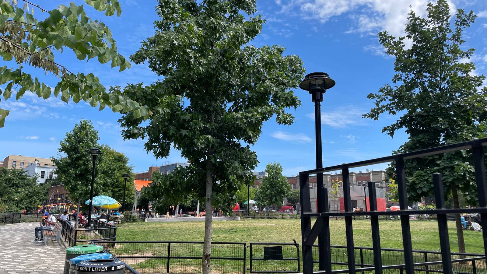Photograph of a small park on a sunny day with young trees, a black metal fence around a grass lawn, a paved path with trash and recycling bins, a tall black lamp post, a blue umbrella and a few people sitting far away, and low brick buildings behind