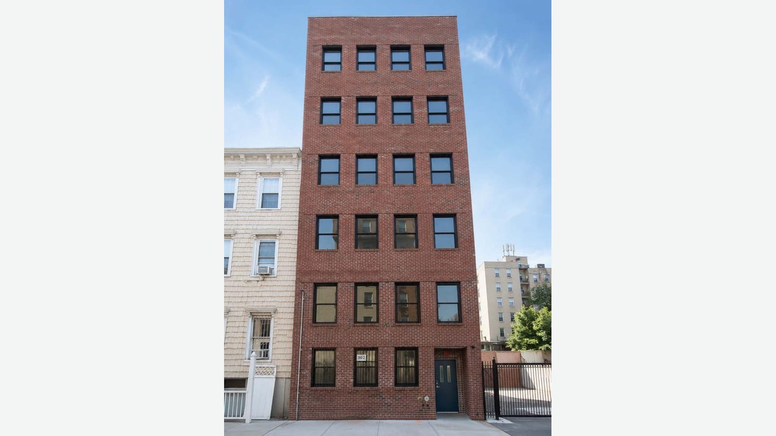 Photograph of a six-story red-brick apartment building with four black-framed windows on each floor, barred windows and a dark blue door on the ground floor, a three-story shingle-sided house to the left and a black metal gate in front of an open lot to the right, with a taller beige apartment building behind, under a blue sky, shown on a plain light background