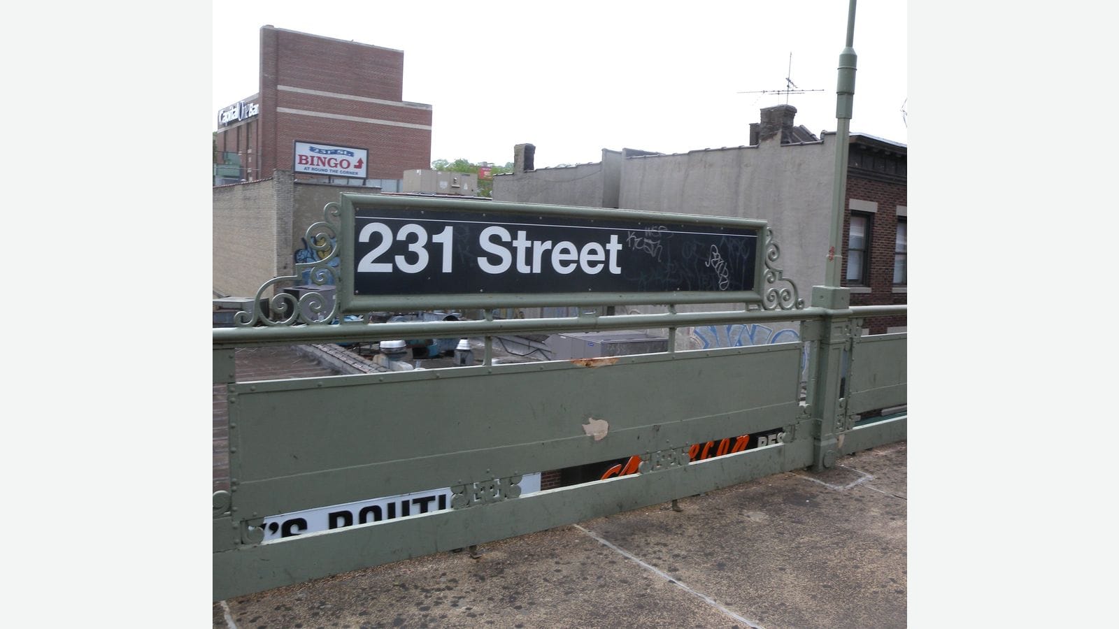 Photograph of a black station sign reading 231 Street in white letters on a green metal railing of an elevated platform, with low brick and stucco buildings, rooftops and a sign for a bingo hall behind, under a white sky, shown on a plain light background