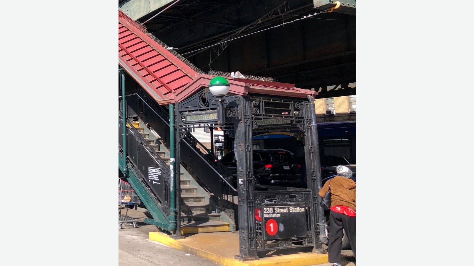 Photograph of a subway entrance under an elevated train line, with a red-roofed green staircase, an ornate black iron entrance frame with a green and white globe lamp and a sign reading 238 Street Station, Manhattan, with a red 1 symbol, and a woman seen from behind at right, shown on a plain light background