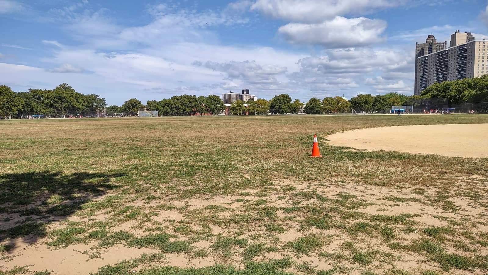 Photograph of a wide, patchy grass ballfield with a sandy infield edge and an orange traffic cone, rows of leafy trees behind, small distant figures on far fields, and tall apartment towers at right under a blue sky with white clouds