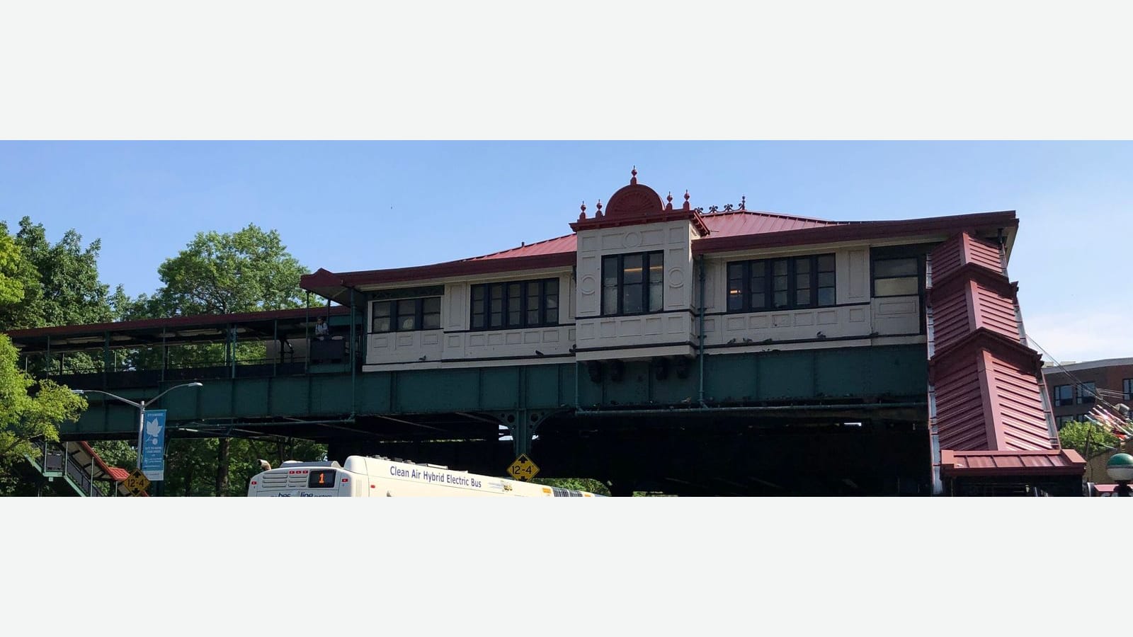 Photograph of an elevated subway station house with cream walls, dark window frames, a red metal roof with a small ornamental crest and a covered red staircase at right, above a green steel structure, with trees at left and the top of a white bus marked 1 below, shown on a plain light background