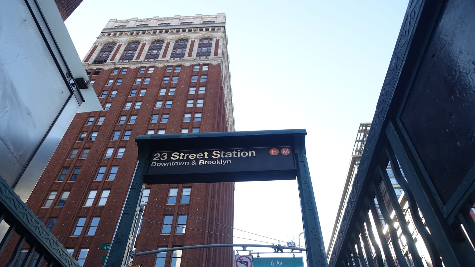 Photograph looking up from a subway stair at a dark green entrance frame with a sign reading 23 Street Station, Downtown and Brooklyn, with F and M symbols, a tall red brick tower with a pale stone top behind it, and a street sign for 6 Av