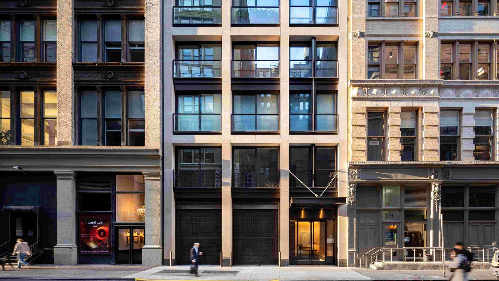 Photograph of a street front with a pale stone grid framing large glass windows, dark railings, two dark metal garage doors and a lit glass entrance under a small canopy, between a brick loft building with a liquor advertisement in its window on the left and a carved stone building numbered 24 on the right, with a man walking past