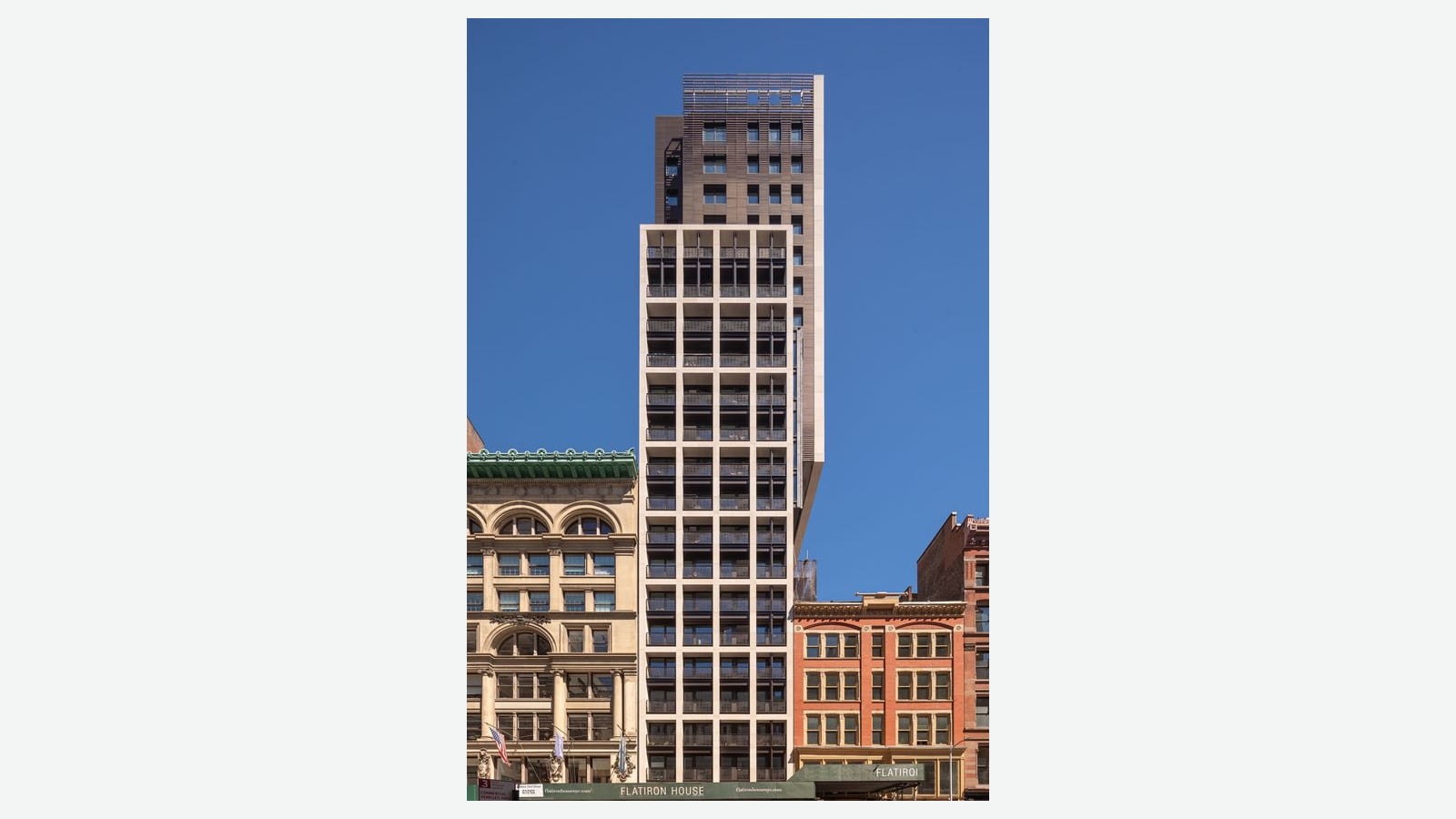 Photograph of a tall, narrow residential tower against a clear blue sky: a lower section with a pale stone grid of square openings and dark balcony railings, a darker brown upper section with small windows, an ornate older building with a green cornice on the left, a red brick loft building on the right and a green sidewalk shed lettered Flatiron House at street level, shown on a plain light background