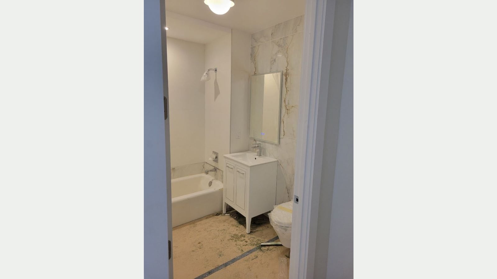 Photograph of a bathroom seen through a doorway, with white wall tile around a white tub and shower head, a white vanity with a sink, a lit mirror on a marble-look tile wall, a toilet still wrapped in plastic, and a bare, dusty floor