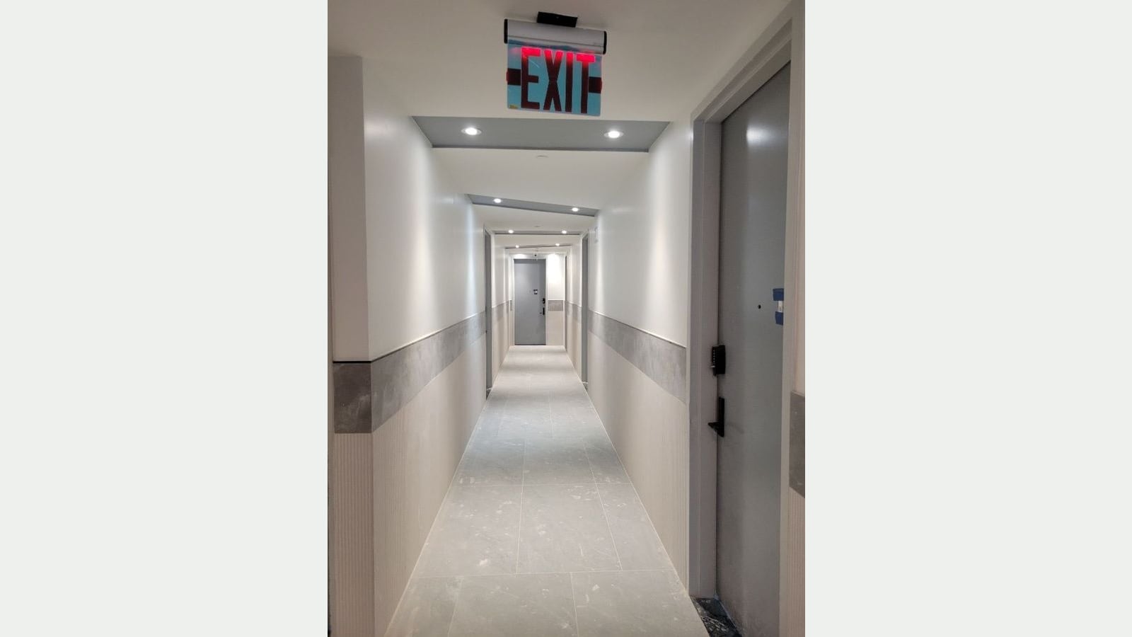 Photograph of a long building hallway with light gray floor tile, white walls with a gray stone band, recessed ceiling lights, gray apartment doors with keypad locks and a lit red EXIT sign overhead