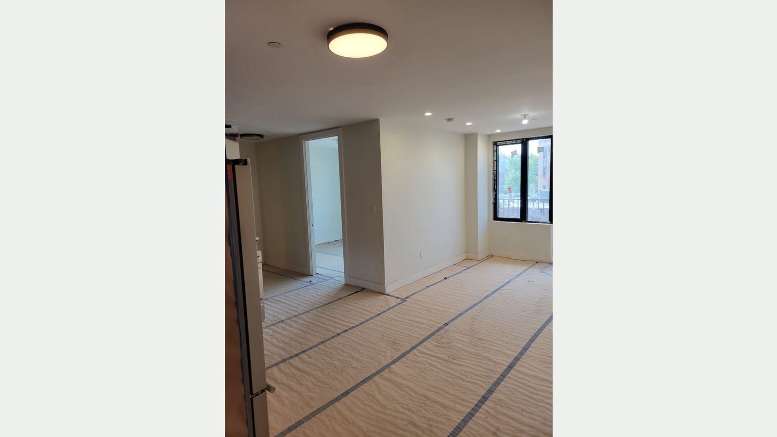 Photograph of an empty room with white walls, a round ceiling light, recessed lights, an open door to a second room, a black-framed window with trees and buildings outside, and protective paper taped over the floor