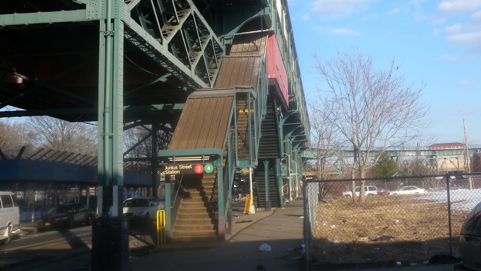 Photograph of a covered street stair climbing to an elevated green steel subway structure, with a sign reading Junius Street Station and red 3 and green 4 symbols, parked cars at left, and a fenced empty lot with bare trees and patches of snow at right
