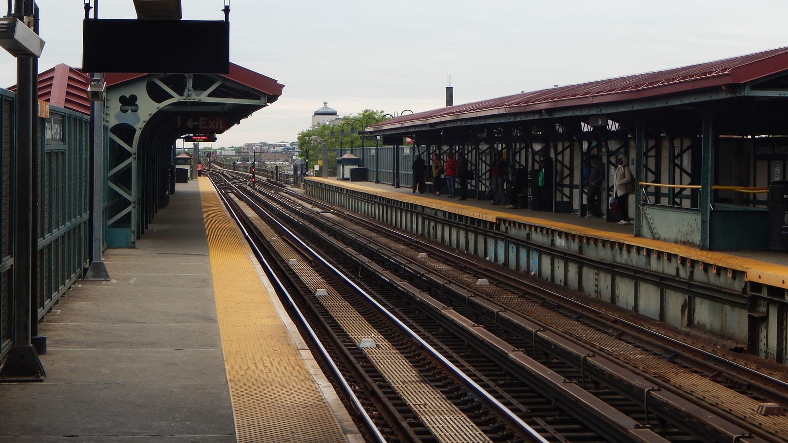 Photograph of an elevated subway station with two platforms under red-roofed canopies on green steel, tracks between them, an Exit sign at left, and a few people waiting far off on the right-hand platform