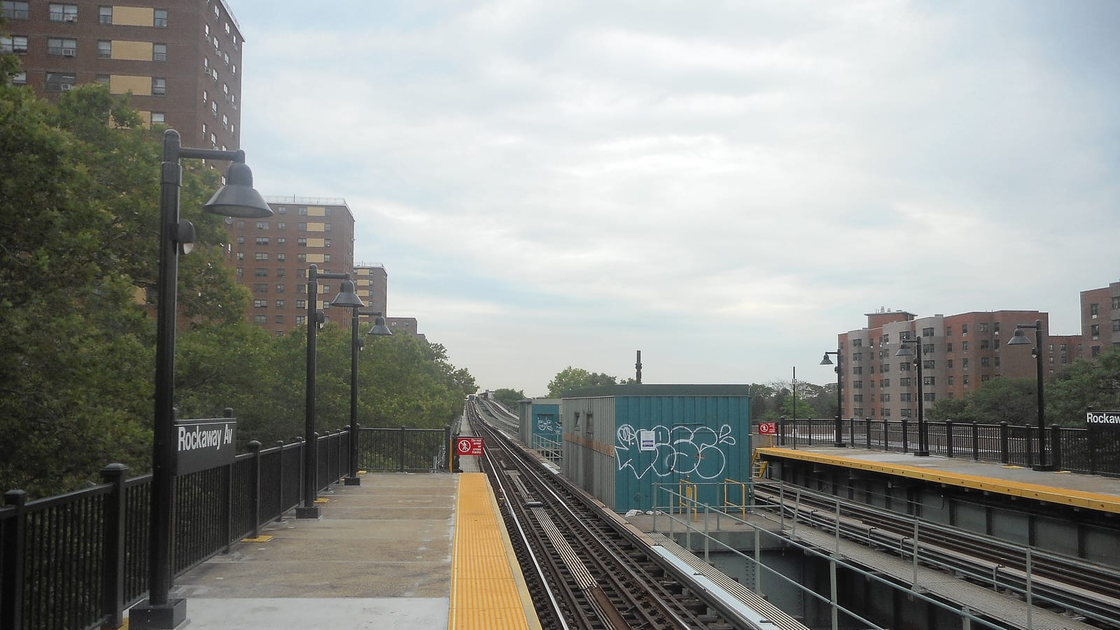 Photograph of an empty elevated subway platform with a yellow edge strip, black lamp posts and a black sign reading Rockaway Av on the railing, tracks running into the distance, and tall brick apartment towers on both sides under a gray sky