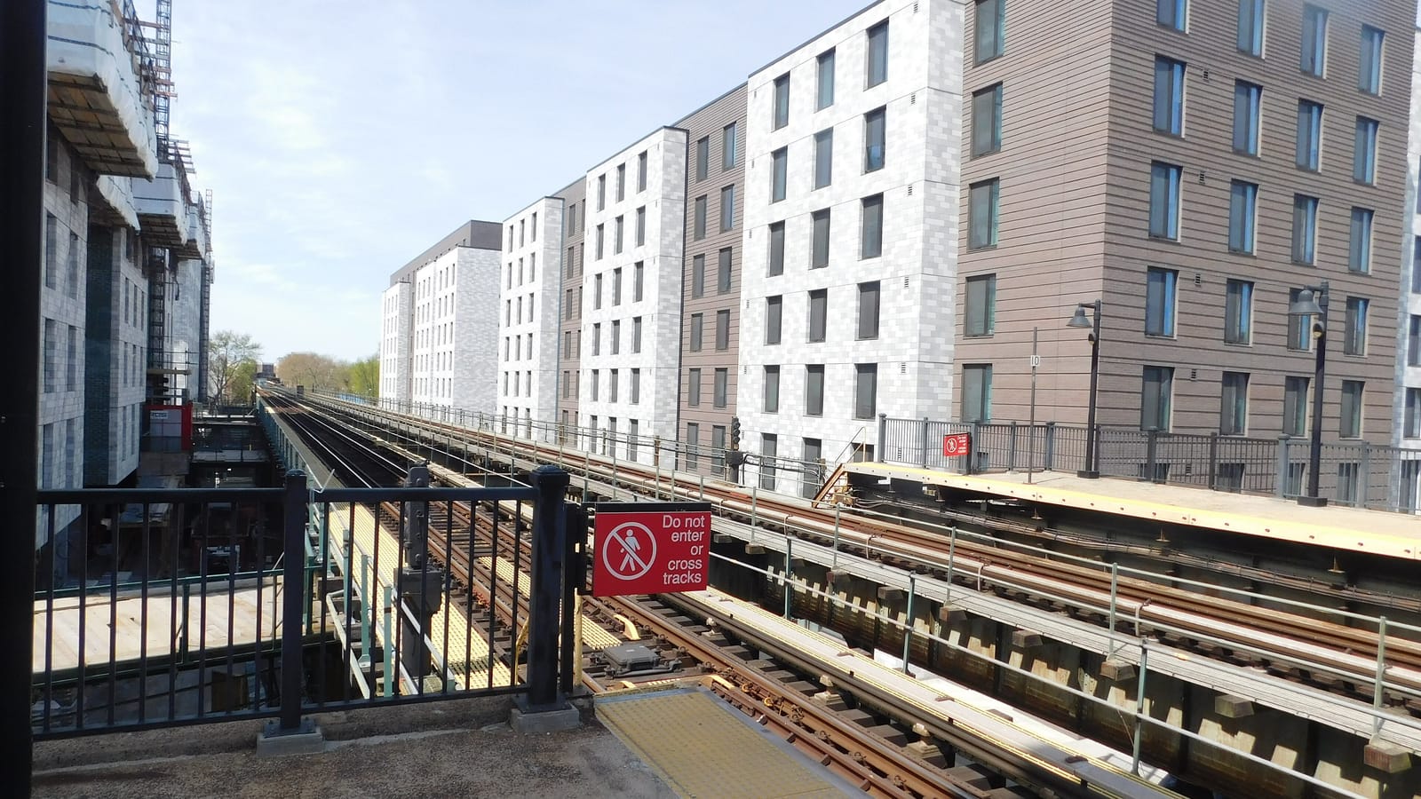 Photograph from an elevated subway platform of tracks curving away, a red sign reading Do not enter or cross tracks, a row of new mid-rise apartment buildings in gray, white and brown beside the tracks, and a building under construction with scaffolding at left