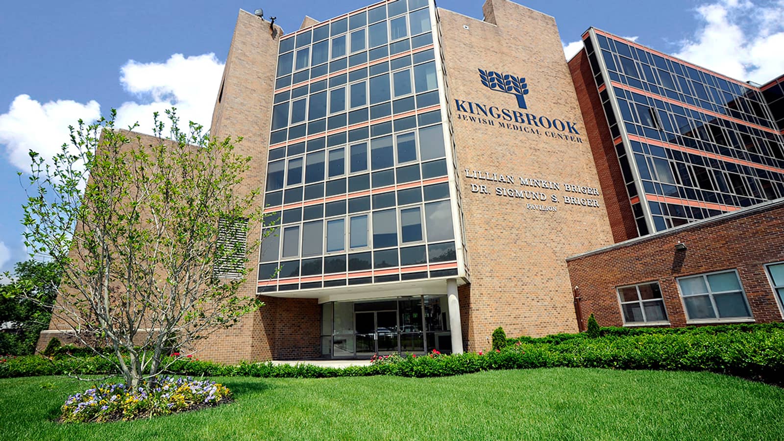 A tan brick hospital building with a tall glass curtain wall, a sign reading Kingsbrook Jewish Medical Center, a young tree and a flower bed on a green lawn in front, under a blue sky with clouds