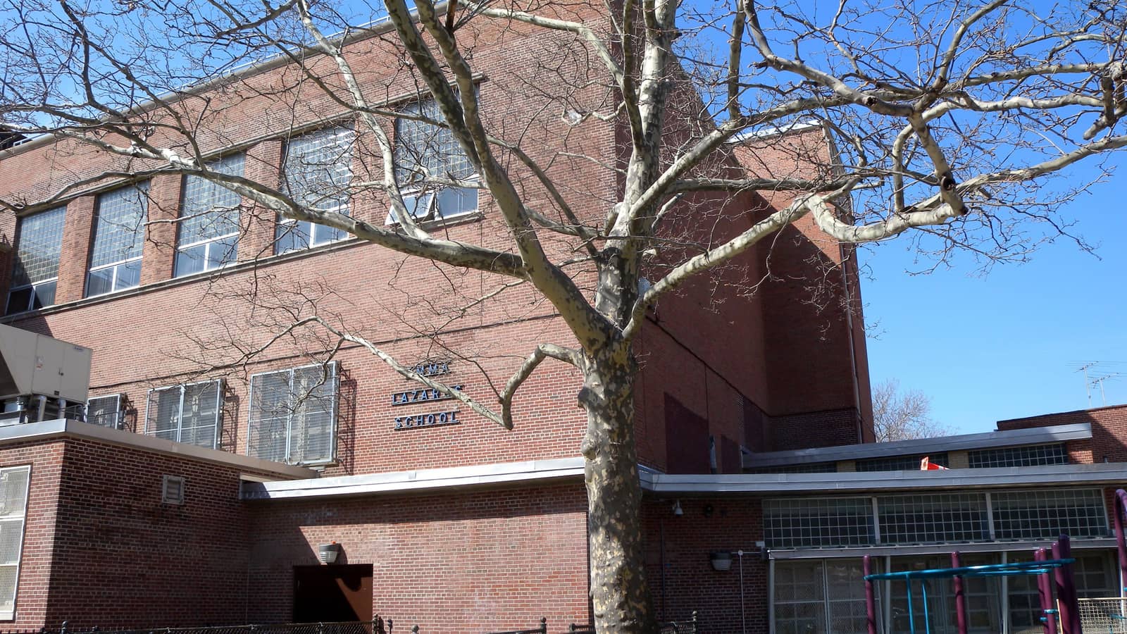 A three-story red brick school building with large steel-framed windows and raised letters reading Emma Lazarus School, behind a bare tree and a black iron fence, with a playground at right, under a blue sky