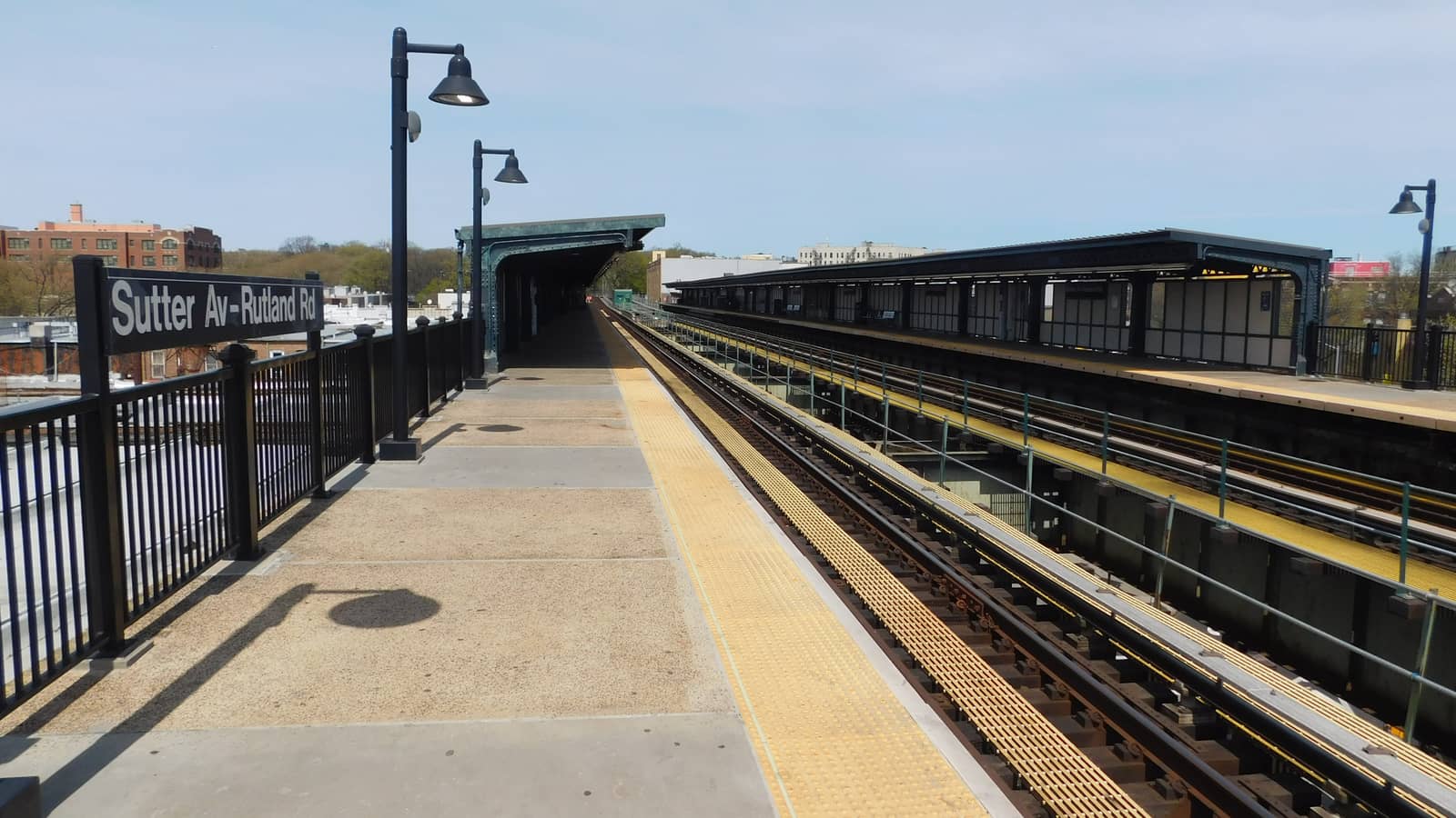 An open elevated subway platform with a black fence, tall black lamp posts, a sign reading Sutter Av-Rutland Rd and tracks running toward covered platforms, with brick apartment buildings beyond