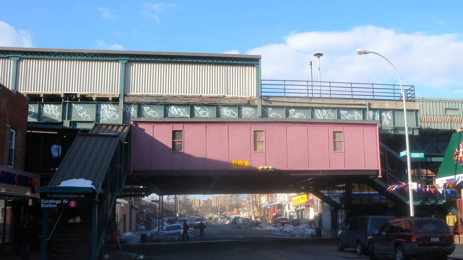 An elevated subway station over a wide street, with a pink enclosed overpass, green steel girders marked with graffiti, a covered stair down to the sidewalk with a sign reading Saratoga Av Station and the number 3, parked cars and leftover snow under a blue sky
