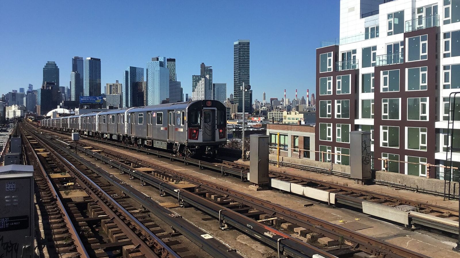 Photograph from the elevated 40 St-Lowery St station looking west along the 7 line: a silver 7 train on the middle track, rusted rails and track equipment in the foreground, the Long Island City and Manhattan skylines in the distance, and a new white and dark-red apartment building at the right edge, under a clear blue sky