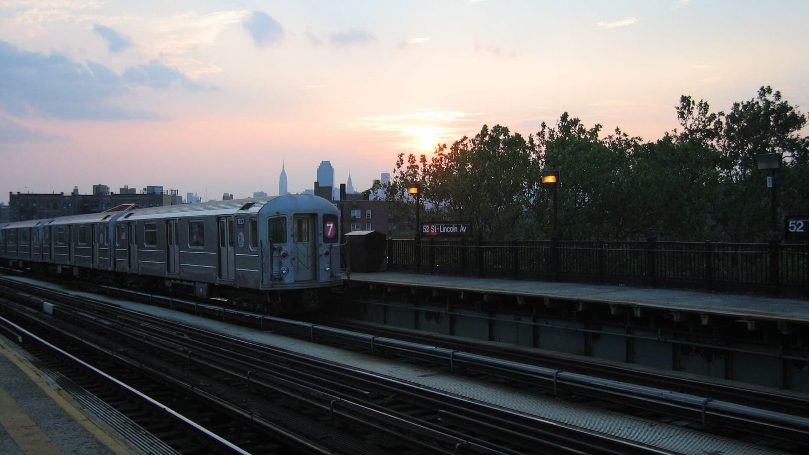 Photograph of an older silver 7 train stopped at the elevated 52 St-Lincoln Av station at sunset, with the station sign, platform lamps and railings at right, trees behind the platform, and the Manhattan skyline with the Empire State Building on the horizon