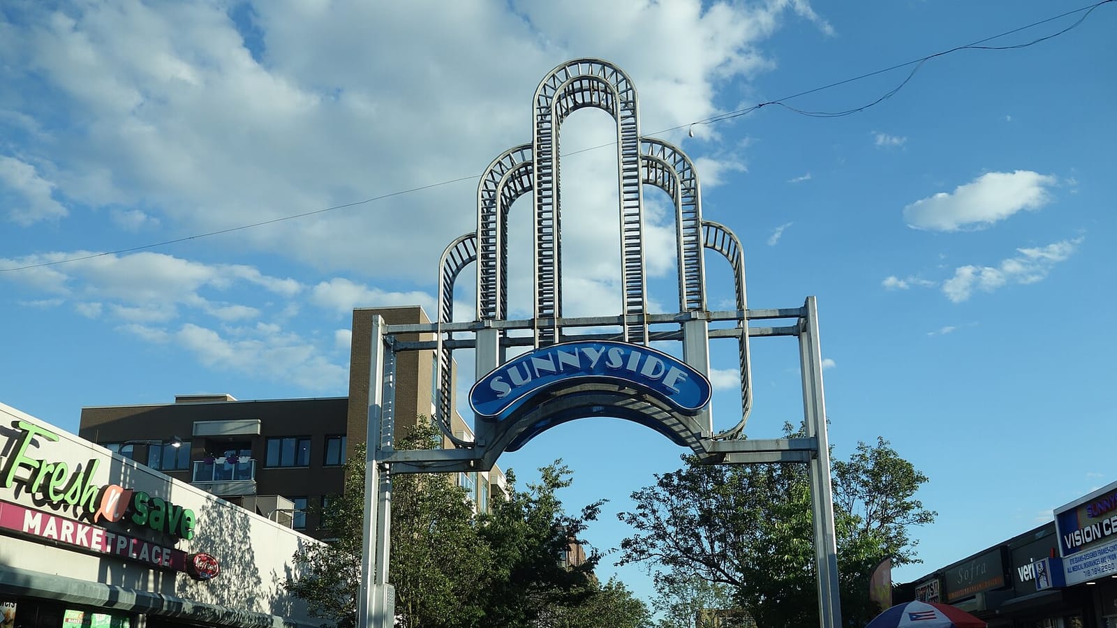 Photograph of the Sunnyside Arch, a tall steel archway with stacked rounded frames and a blue sign reading 'Sunnyside', over a street lined with trees, a supermarket front on the left, shops on the right and a brown brick apartment building behind, under a blue sky with clouds