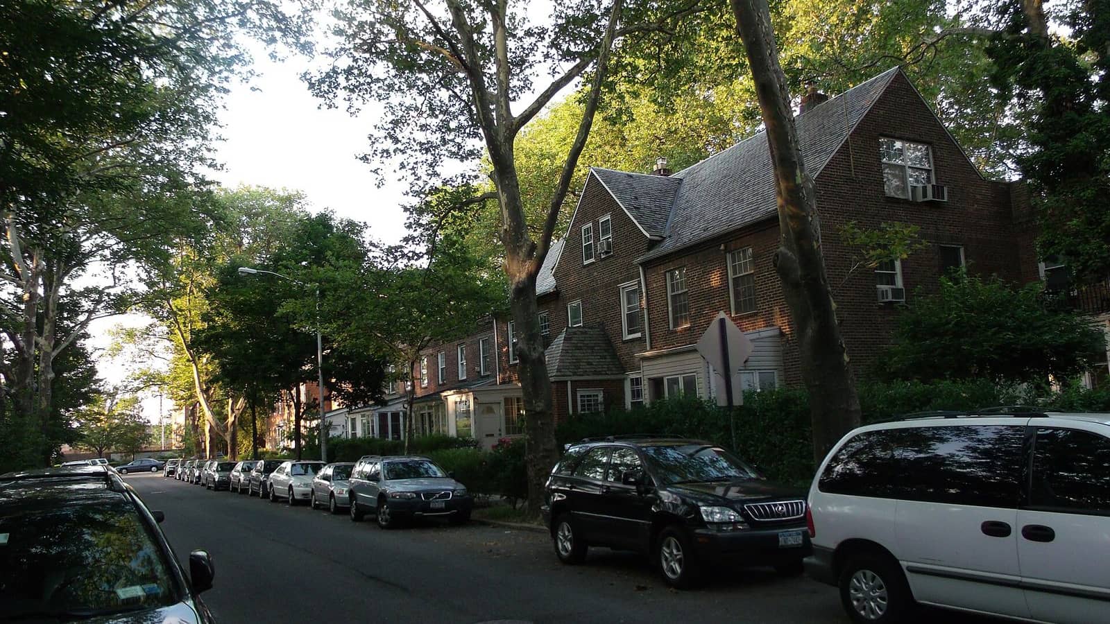 Photograph of a shaded residential street in the Sunnyside Gardens Historic District, with two- and three-story brick row houses with steep slate roofs and small front gardens behind hedges, tall plane trees, and cars parked along both curbs