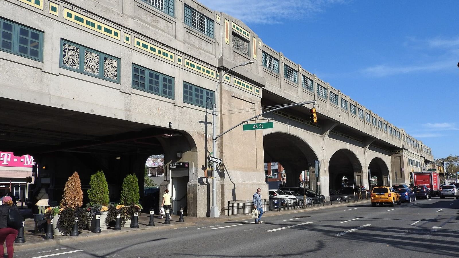 Photograph of the elevated 46 St-Bliss St subway station in Sunnyside, a long gray concrete structure with green-framed windows, yellow and green tile bands and arches over the street, a green '46 St' street sign on a traffic-light pole, a station entrance sign reading '46 St-Bliss St Station' with a 7 bullet, planters and bollards on the sidewalk, a few people walking and a yellow taxi on the road under a blue sky
