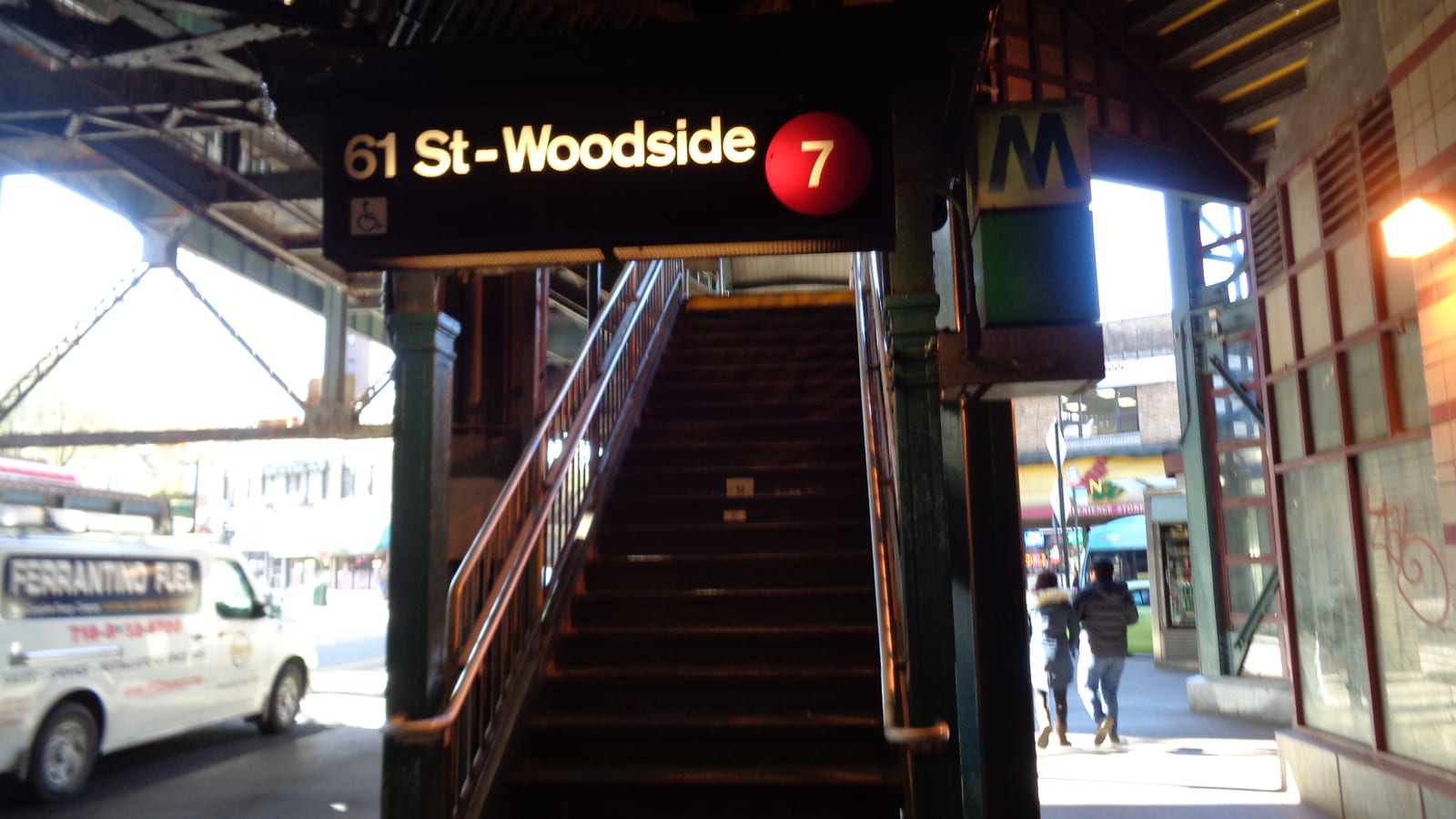 A lit black sign reading 61 St-Woodside with a red 7 circle and a wheelchair symbol over a steep stairway under steel train tracks, a white fuel delivery van at left and two people seen from behind walking along the sidewalk at right