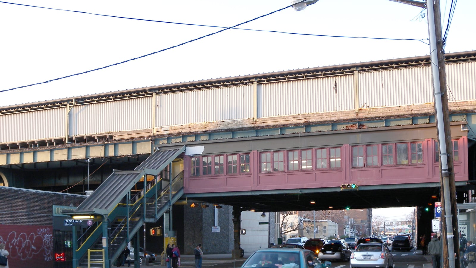 A pink station house with rows of small windows hanging under the steel structure of a train line that crosses a street, with a covered stairway at left beside a sign reading 69 St-Fisk Av Station and a 7 symbol, cars in traffic below and people waiting at the corner